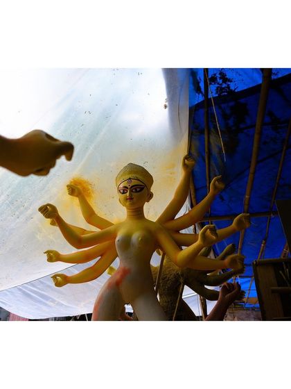 An unfinished Durga idol stands under a blue tarp, creating a dramatic silhouette. This image captures the anticipation before the final reveal.