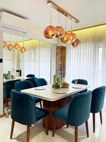 A luxurious dining setup featuring deep teal velvet chairs and a statement cluster of rose-gold pendant lights. The large mirror wall doubles the space and adds a layer of glamour.