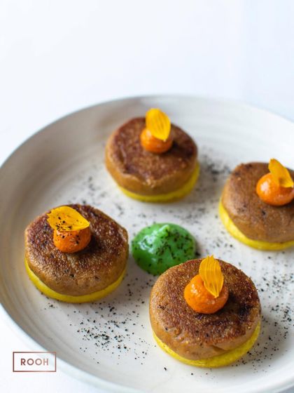 Four Raw Banana and Fig Galouti kebabs, elegantly plated with a green chutney and garnished with a candied fruit.