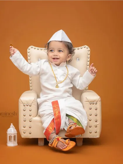 A cheerful little boy in a traditional white dhoti and Gandhi cap, seated on a miniature throne. This simple setup creates a distinguished and cute portrait.