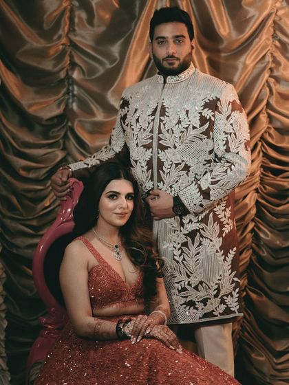 A formal yet intimate couple portrait from their sangeet. The bride is seated while the groom stands beside her, their coordinated outfits and regal poses creating a picture of pure elegance.