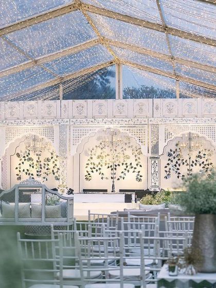 A view of the sangeet stage from a distance, framed by foliage. The scale and beauty of the Thikri backdrop are evident even from afar.