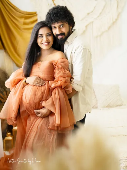 A beautiful portrait of a smiling couple in a boho-chic setting. The expecting mother in a peach gown and her partner pose together, radiating happiness.