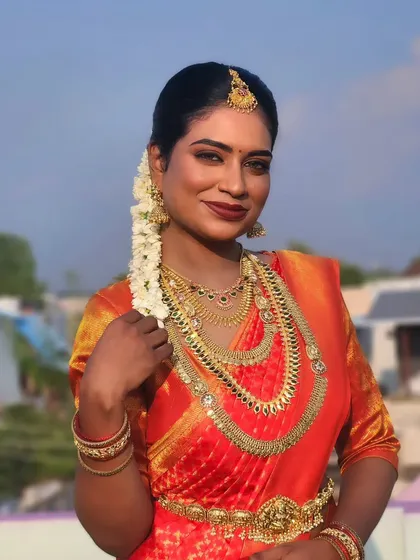 A stunning portrait of a dusky bride against a blue sky. The makeup is kept natural and glowing, with a bold red lip that complements her beautiful skin tone and orange saree.
