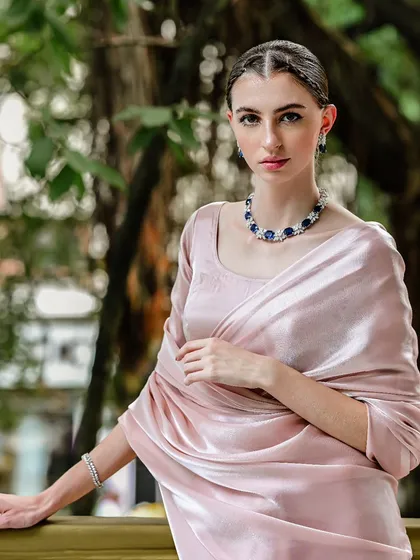 A jewellery campaign shot featuring a model in a delicate pink satin saree. The outdoor setting with natural light beautifully illuminates the sapphire necklace.