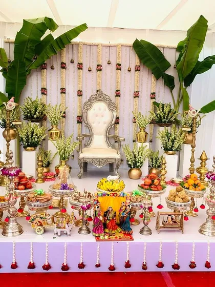 An elegant and bright Seemantha decoration featuring a white backdrop with hanging sugandharaja garlands. The stage is flanked by banana plants and a symmetrical arrangement of offerings, creating a pure and sacred ambiance.