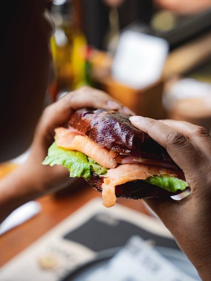 A close-up of hands holding a lye-dipped bagel sandwich, showing the rich color of the crust and the fresh fillings inside.
