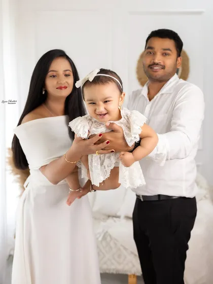 A proud family holding their smiling baby girl. Her lacy dress and bow complement the bright, airy feel of the studio setting.