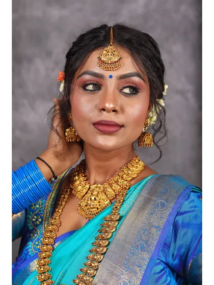 A thoughtful portrait of a model in a blue silk saree. The focus is on her expression and the stunning temple jewelry, including the necklace and maang tikka.