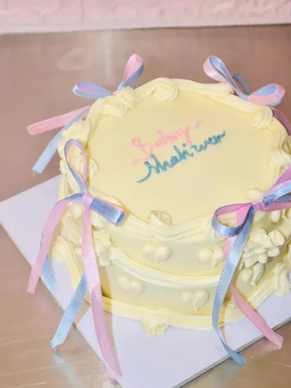 We love the delicate, vintage coquette cake trend. This beautiful baby shower cake features soft yellow buttercream, intricate piping, and is adorned with delicate pink and blue ribbons, making it a perfect centerpiece.