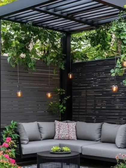 This design features a sleek, black pergola with climbing vines, creating a natural canopy over a comfortable L-shaped sofa. The dark, slatted privacy screen and lush surrounding greenery provide a sense of seclusion and tranquility.