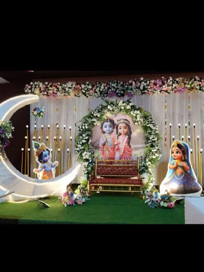 A divine naming ceremony stage with a Krishna theme. The setup features a decorated jhula, a large moon prop, and cutouts of little Krishna and Radha.