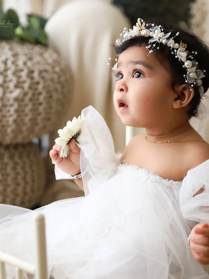 A baby girl in a white tulle dress and pearl headband looks up with wonder, holding a flower.