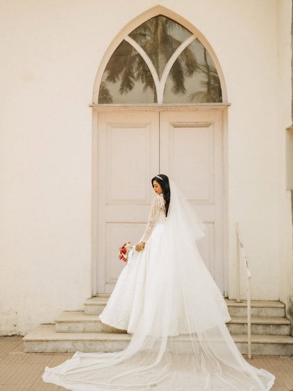 An elegant bridal portrait showing the full length of the bride's gown and veil. The simple white architecture of the church provides a perfect backdrop.