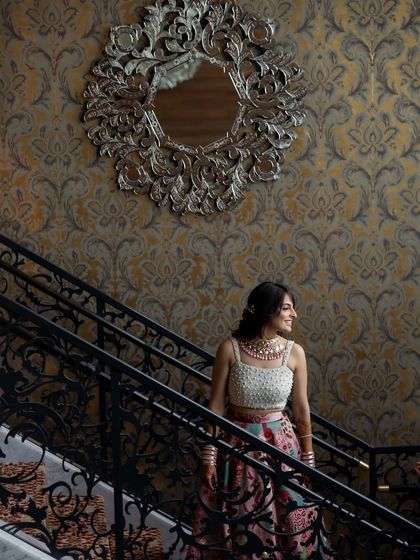 A bride poses on a grand staircase, her elegant lehenga and jewelry perfectly complemented by the ornate surroundings.