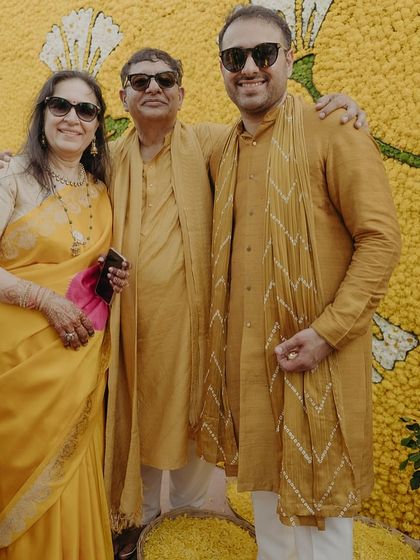 The family posing against a backdrop of yellow flowers at the Haldi ceremony. The parents and groom are styled in coordinated shades of mustard yellow, creating a warm and cohesive family portrait.