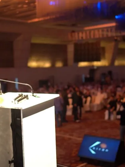 Addressing the audience at the Kiran Gems event, with the company's branding visible on the lectern and screen.