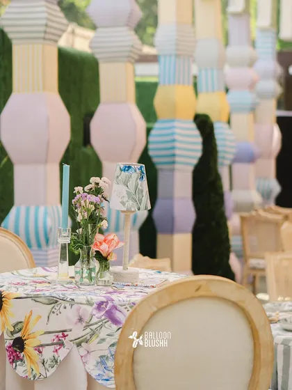 A table setting with a floral tablecloth, a small matching lamp, and a backdrop of custom-built pastel-colored pillars.