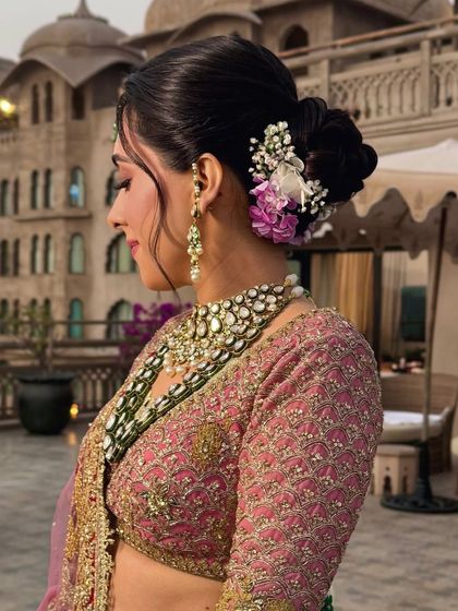 A soft, romantic updo with wispy tendrils, perfect for a sunset wedding. The bun is accented with delicate purple and white flowers for a touch of color.