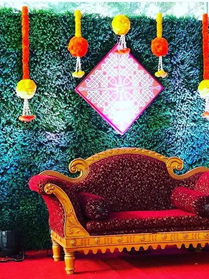 This traditional baby shower backdrop combines a vibrant green wall with hanging marigold garlands and classic brass lamps (diyas). The ornate red sofa provides a royal seating arrangement for the mother-to-be.