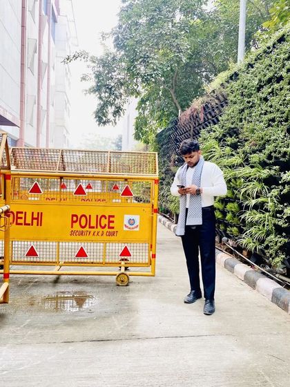 Standing next to a Delhi Police barricade, a familiar sight in my line of work. It represents the boundary between the state and the individual, where I operate to protect my client.