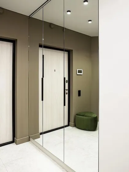 A minimalist entryway featuring a full-length mirrored wall that visually doubles the space. A small green ottoman adds a pop of color and a convenient place to sit.