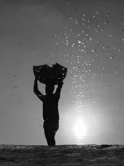 A reflection of a devotee holding an offering during Chhath Puja, captured in a puddle. This black and white shot creates a powerful silhouette against the sun.