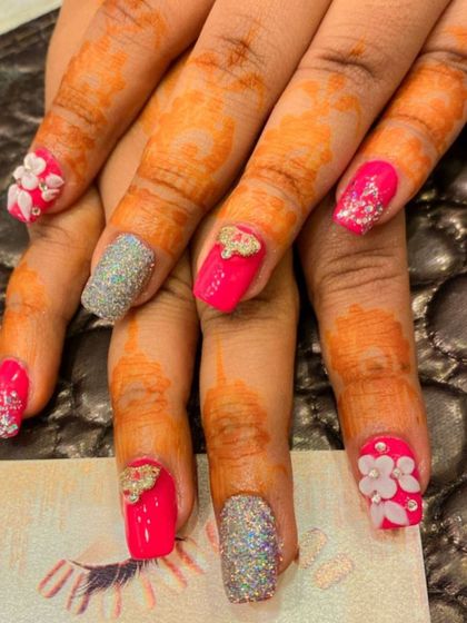 A vibrant and celebratory set with henna-stained hands. The nails feature hot pink polish, silver glitter accents, and 3D elements like bows and flowers.