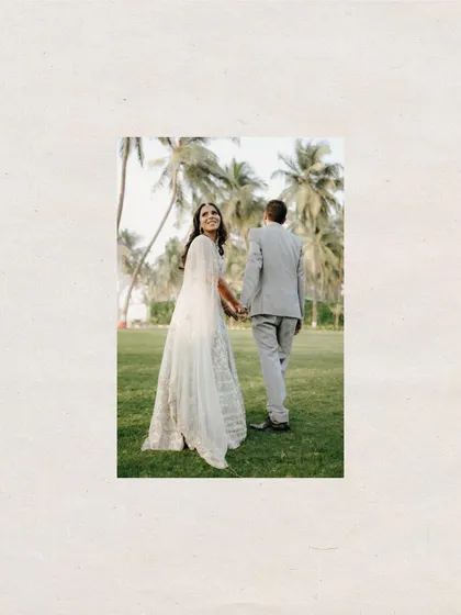 A classic "follow me to" style shot, framed beautifully. The bride leads the groom through a green lawn, looking back with a smile, symbolizing the start of their journey.