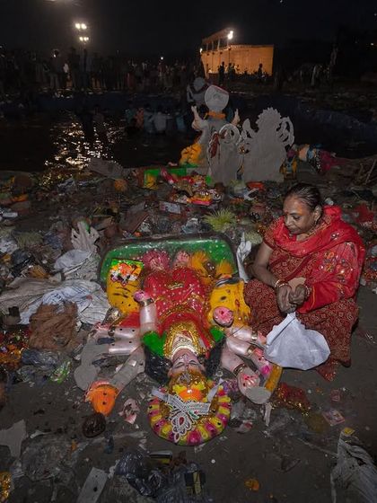 A woman prays over a goddess idol lying on the polluted riverbank after the Visarjan, a somber image reflecting on faith and environmental impact.