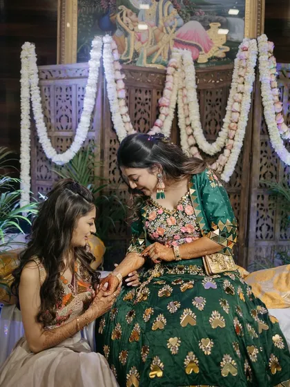 The decor should be the perfect backdrop for candid moments like this one. The traditional wooden screen and fresh flower garlands create a rich, textured background that enhances the beauty of the Mehendi ceremony.