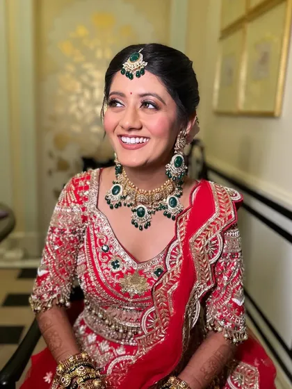 A joyful bride with a natural and radiant makeup look. The soft eyeshadow and glowing skin perfectly complement her traditional red lehenga and emerald jewellery.