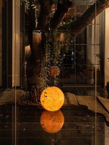 A night shot of the KEUS courtyard, where a glowing orb light sits at the base of a mature tree, reflected in the dark, polished floor. The scene is a serene blend of stone, light, and life.