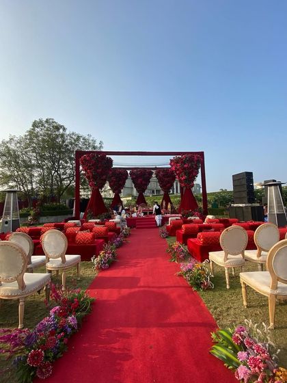 A bold and regal mandap designed entirely in shades of red. The structure features massive floral arrangements resembling trees of roses, creating a powerful and passionate setting for an outdoor wedding ceremony. The long red aisle adds to the dramatic effect.