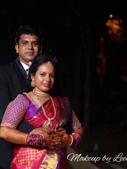 A classic reception portrait of a couple. The bride's makeup features a soft smokey eye and a pink lip, complementing her rich pink and purple silk saree.