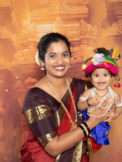A happy portrait of a mother holding her smiling baby, who is dressed in a colorful Krishna costume.
