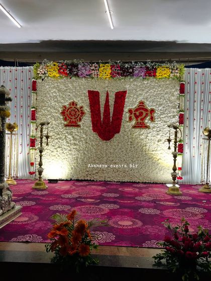 A grand naming ceremony stage with a backdrop of white flowers featuring a large 'Namam' symbol. The setup is completed with traditional brass lamps for a spiritual ambiance.