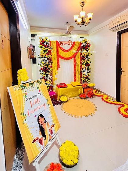 A complete view of a Haldi setup in a home corridor. The decor features a welcome sign, a floral and marigold backdrop, and a flower-lined pathway.
