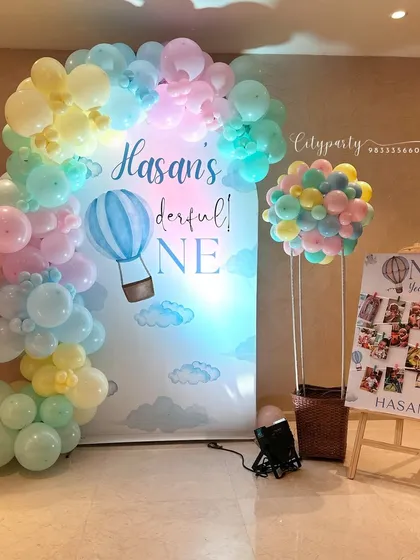This 'Onederful' hot air balloon theme for a first birthday uses a soft pastel color palette. The setup includes a custom backdrop, a neon '1' sign, and a balloon sculpture shaped like a hot air balloon basket.