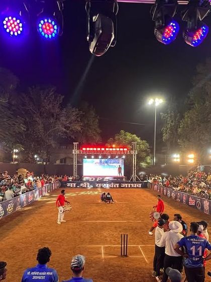 The view from behind the batsman during a night match. You can see the crowd, the powerful overhead lights on the truss, and the main stage with the LED screen, creating a professional tournament atmosphere.