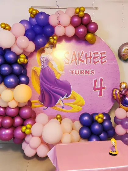 A Rapunzel-themed fourth birthday party featuring a custom circular backdrop with the princess herself. The scene is framed by an organic balloon garland in shades of purple, pink, and gold, matching Rapunzel's dress and hair.