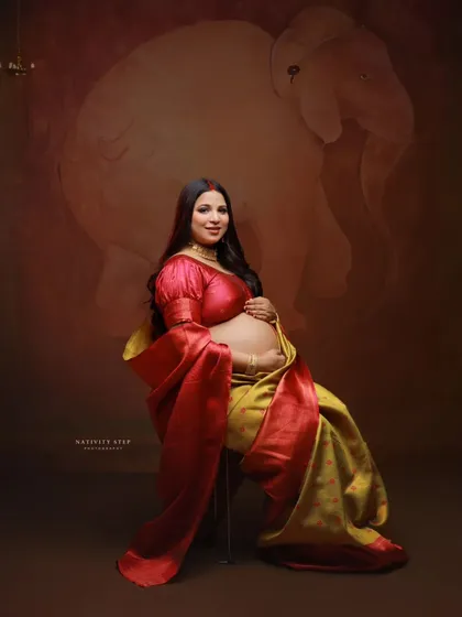 A solo portrait of the mother-to-be seated, showcasing her baby bump in a beautiful yellow and red saree. The pose is both regal and gentle.
