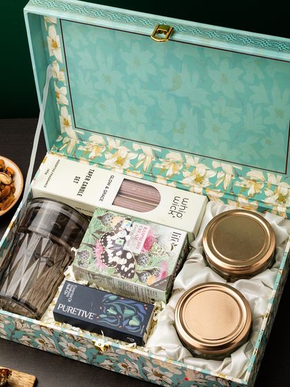 This elegant hamper is presented in a beautiful floral-printed trunk box. Inside, it holds a cut-glass tumbler, taper candles, and other premium items, perfect for a luxury gifting experience.
