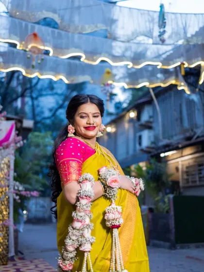 The mother-to-be smiles radiantly, standing under festive outdoor decorations for her baby shower. This shot captures her joy and the celebratory atmosphere of the event, highlighting her traditional attire and floral ornaments.
