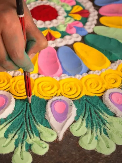 A close-up of a hand meticulously creating a powdered rangoli, using a pencil to refine the edges of the colorful peacock feather design.