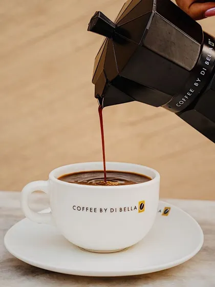 This action shot captures the moment of pouring fresh coffee from a moka pot. The simple, clean setup with a branded cup keeps the focus on the rich, dark liquid, evoking the aroma and taste of a fresh brew.