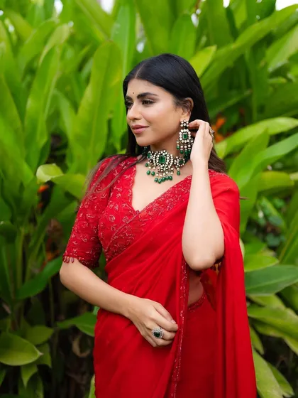 A close-up portrait of a model in a red saree, adjusting her earring. The shot highlights the intricate jewelry and her elegant profile.