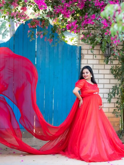 A dramatic shot of a mom-to-be in a red gown against a bright blue gate with pink flowers.