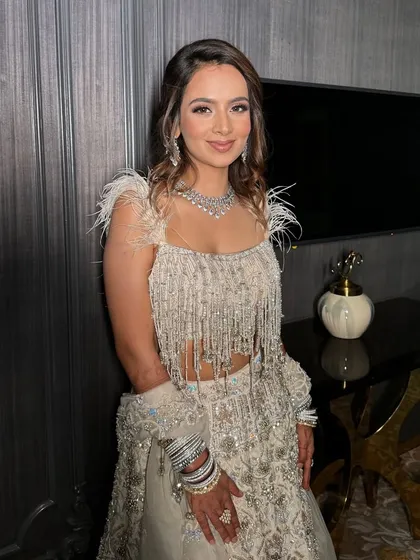 The sister of the groom, shining bright in a feathered and fringed lehenga. Her makeup is soft and glamorous, with a focus on radiant skin and bright eyes.