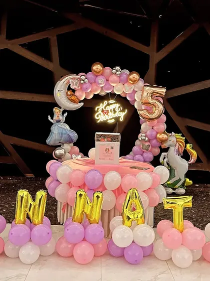 A whimsical princess party for a 5th birthday. The setup includes Cinderella and unicorn props, a crescent moon foil balloon, and balloon lettering spelling out the name 'UNNATI'.